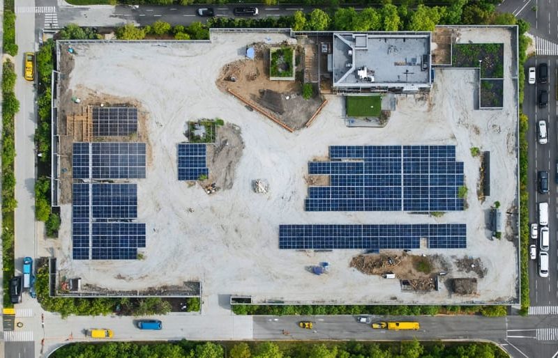 Aerial view of a sustainable construction site featuring solar panels, green roofs, and native landscaping, situated near green spaces and public transit.